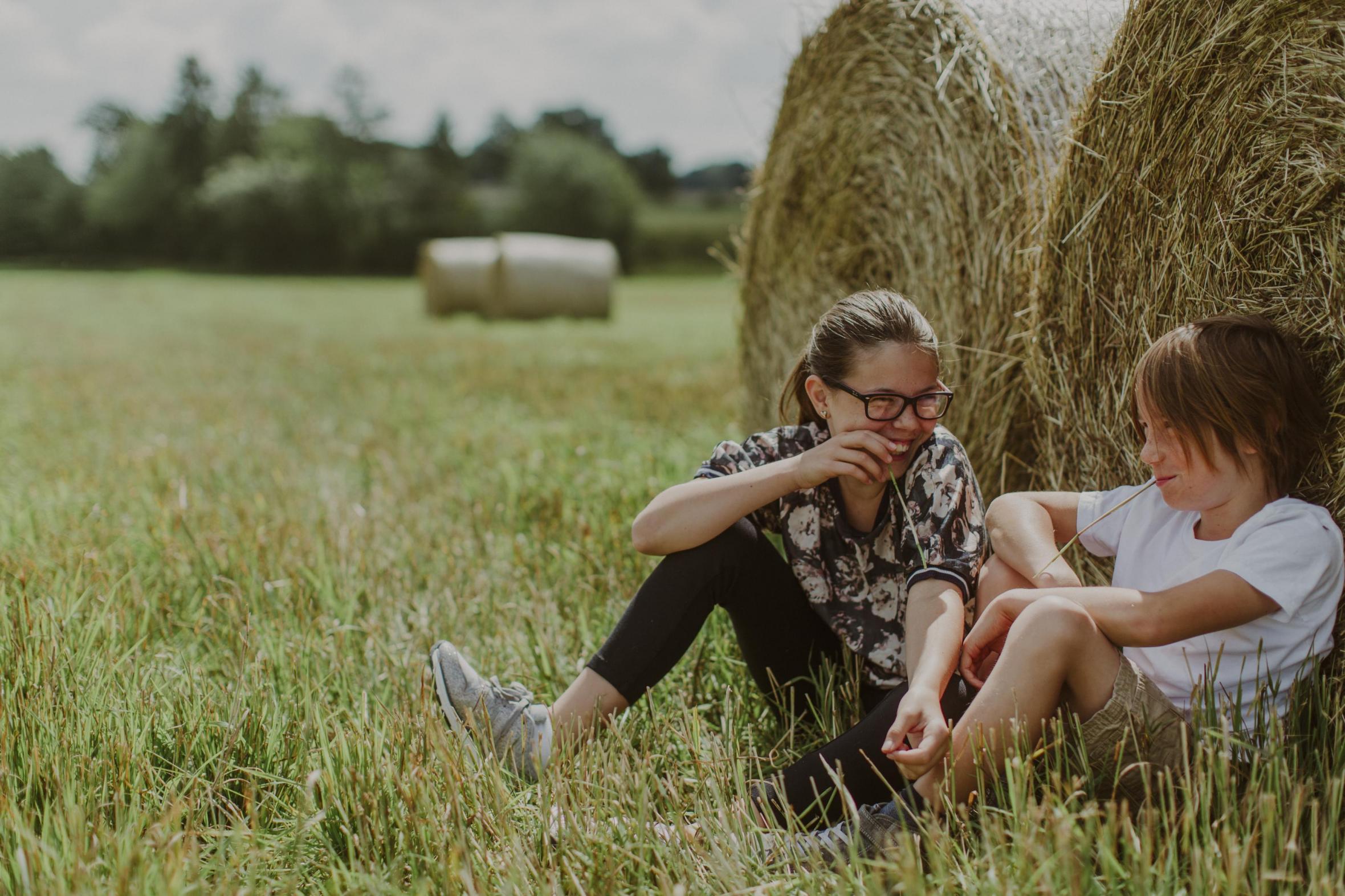 Två barn sitter vid stora höbalar och tar det lugnt på fältet