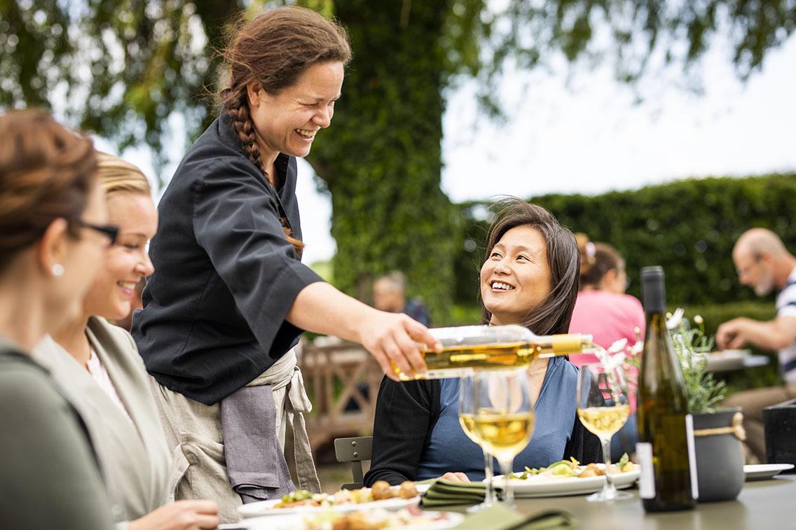 Laughing waitress in black shirt serving three sitting women white wine from a bottle