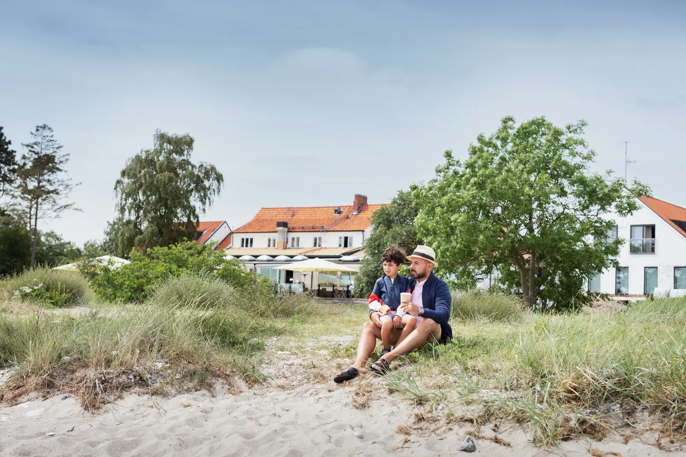 Father sits with his son in front of a white big house at a sandy beach
