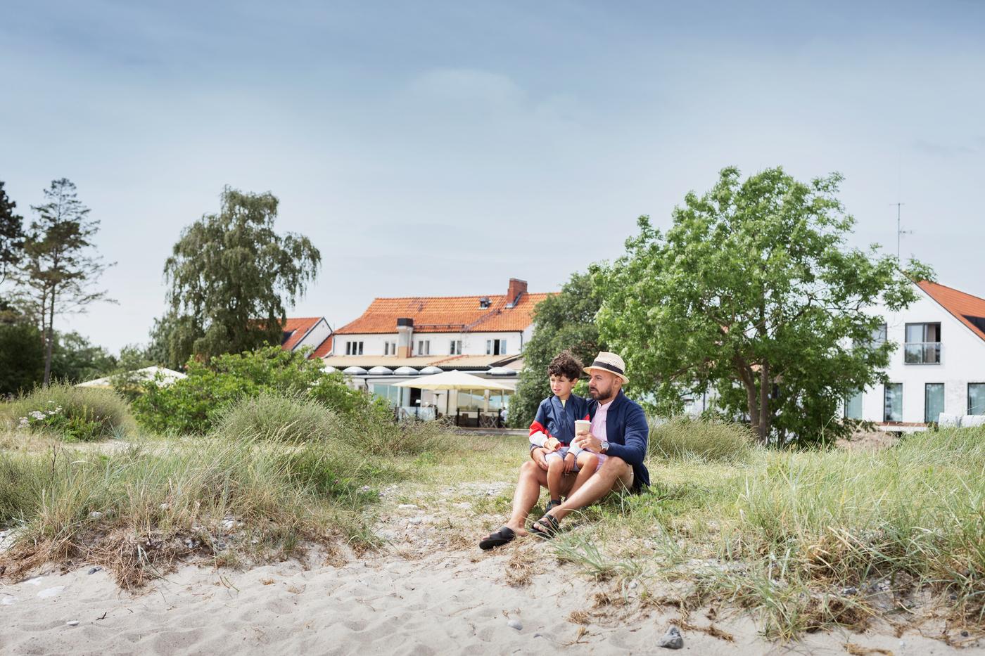 Pappa sitter med sin son sitter framför ett stort vitt hus vid kanten av en sandstrand