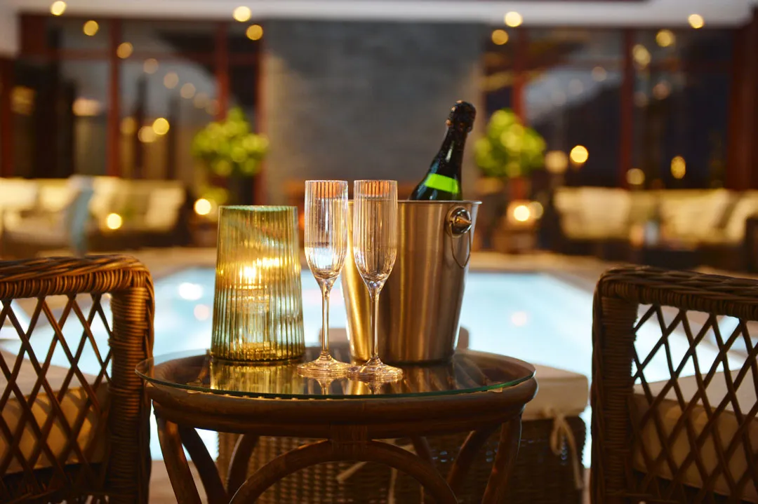 Two chairs and a table with flute glasses and a bottle in front of a pool