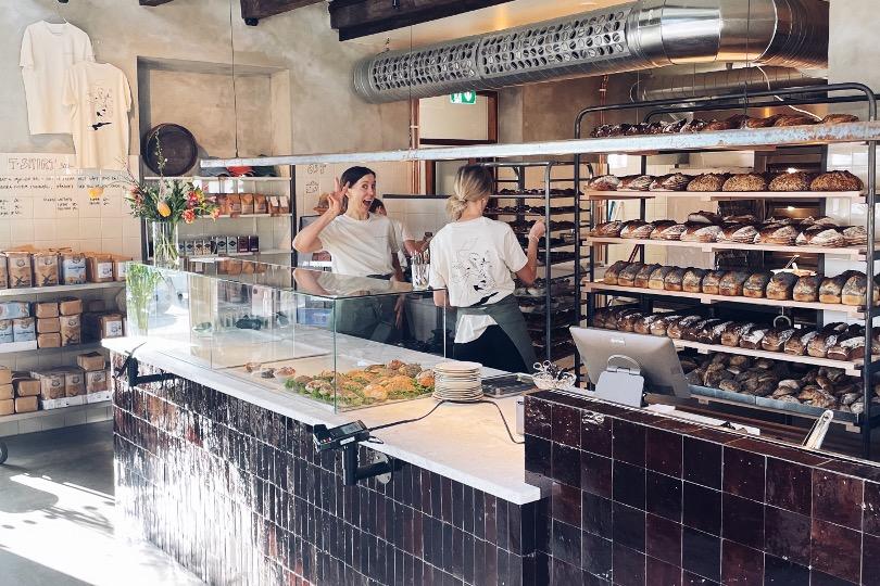 Två glada kvinnor står bakom disken på ett bageri med nybakat bröd vid sidan om sig
