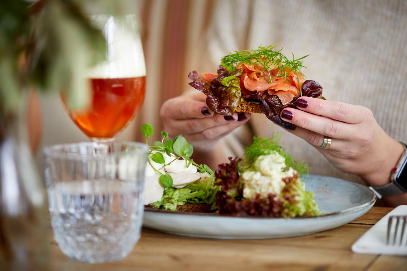 Woman holding generously topped smørrebrød with salmon and garnishes while enjoying a beer