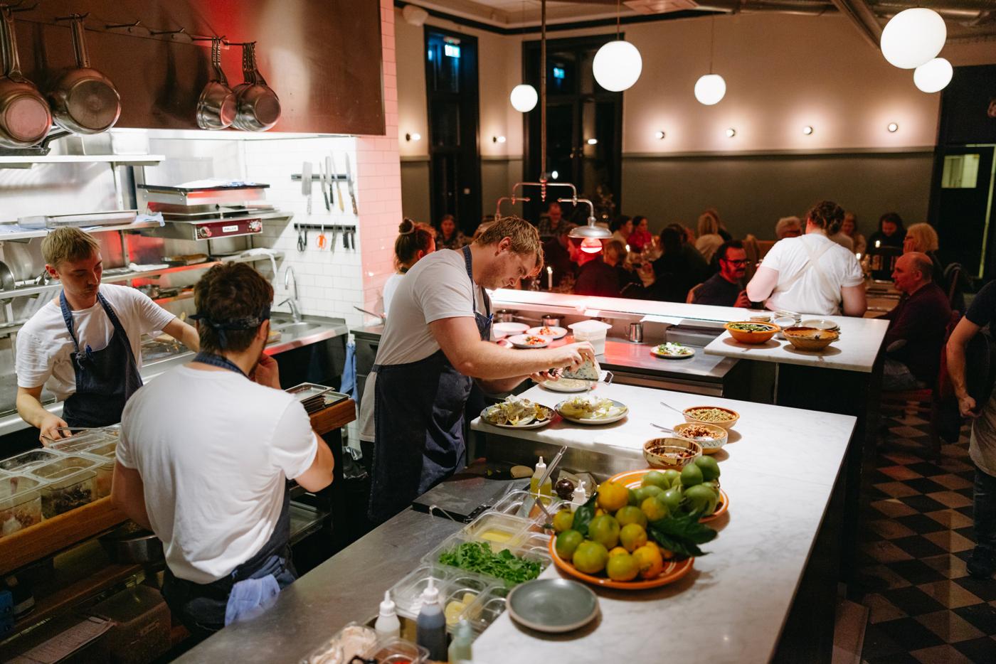 Open kitchen with chefs at work in foreground, dining area with seated guests visible in dimmer background setting