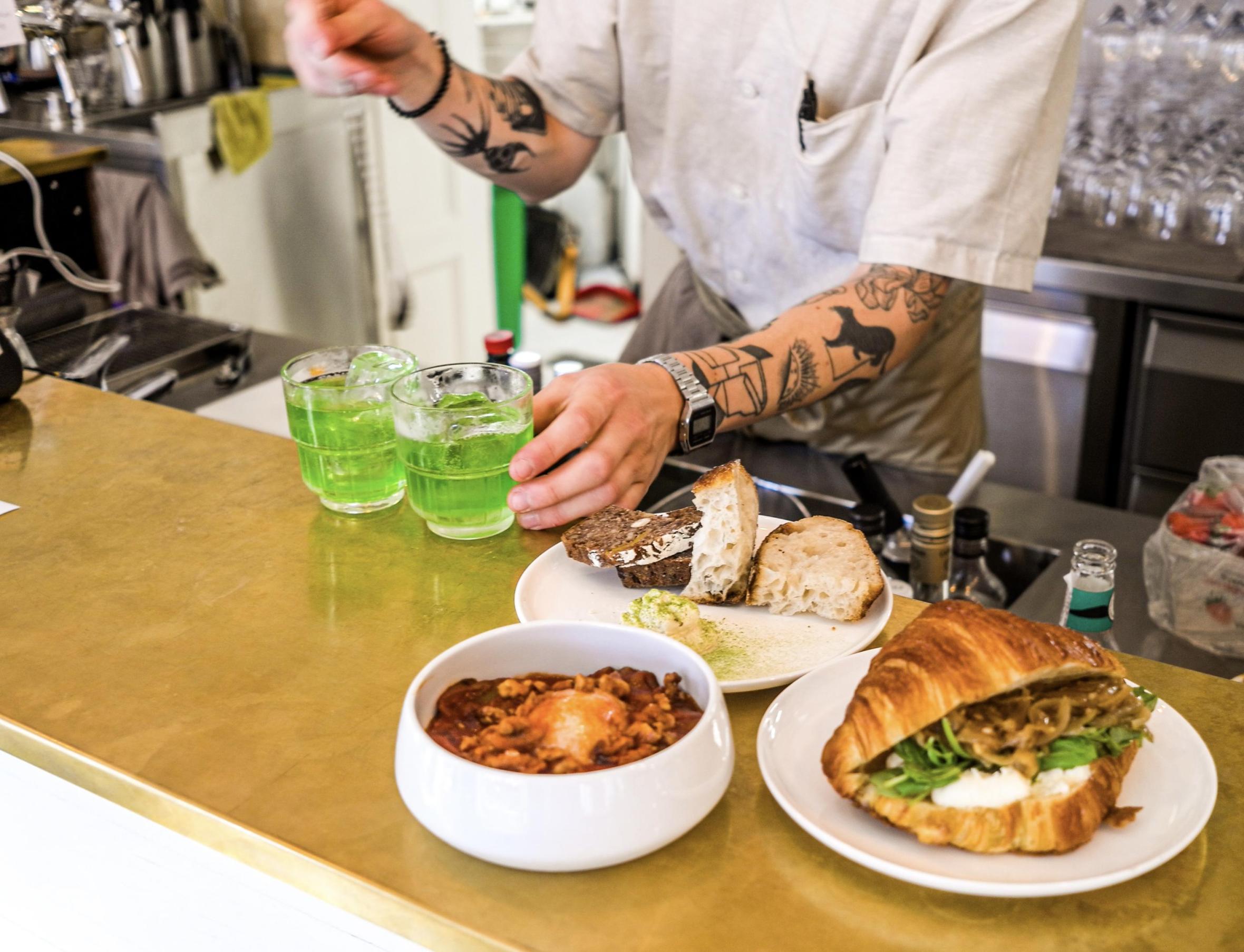 Two cocktails and small plates arranged on bar counter with bartender visible in background preparing drinks