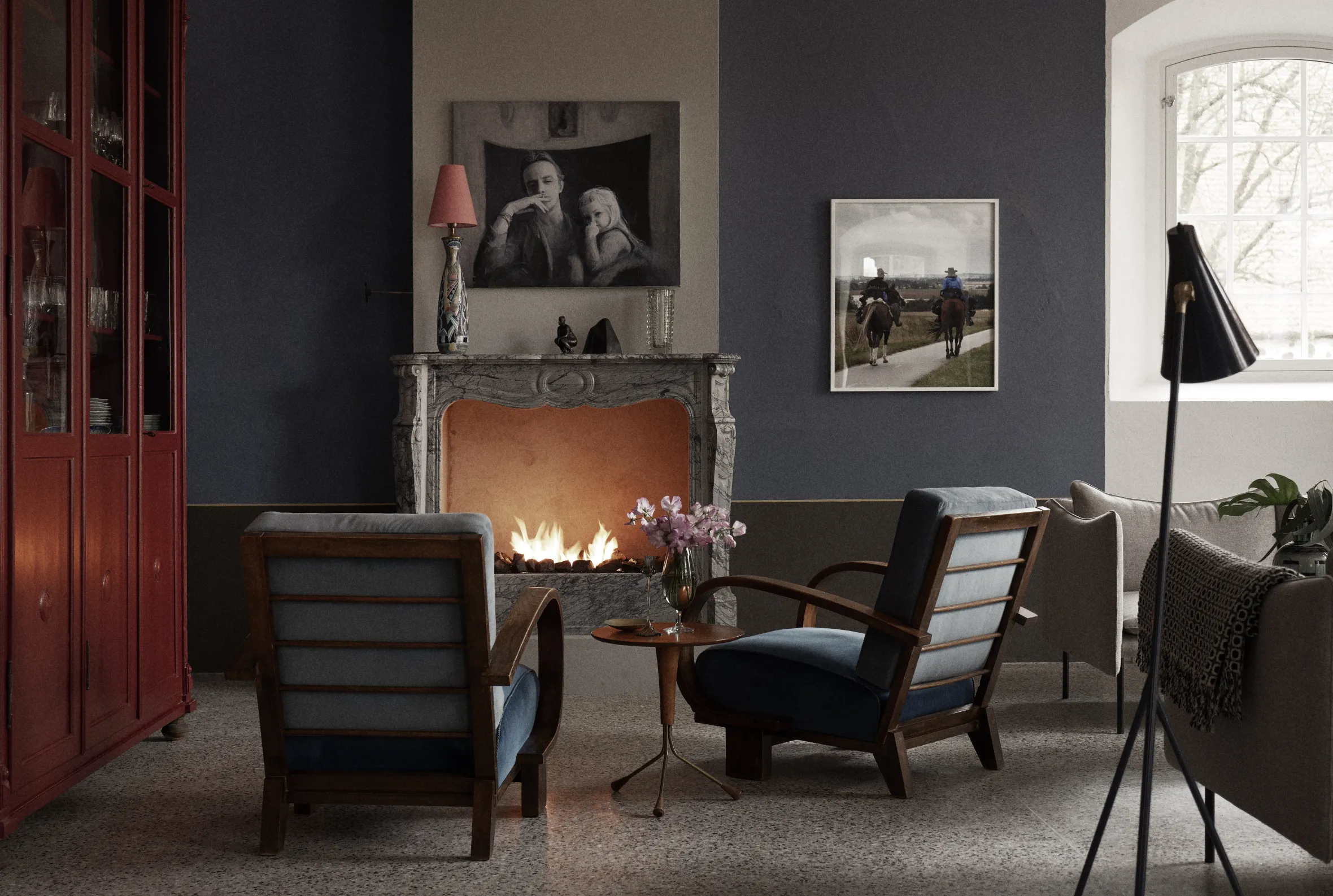 Elegant living room with lit marble fireplace, mid-century armchairs, framed photographs, display cabinet, and standing lamp creating warm, sophisticated atmosphere