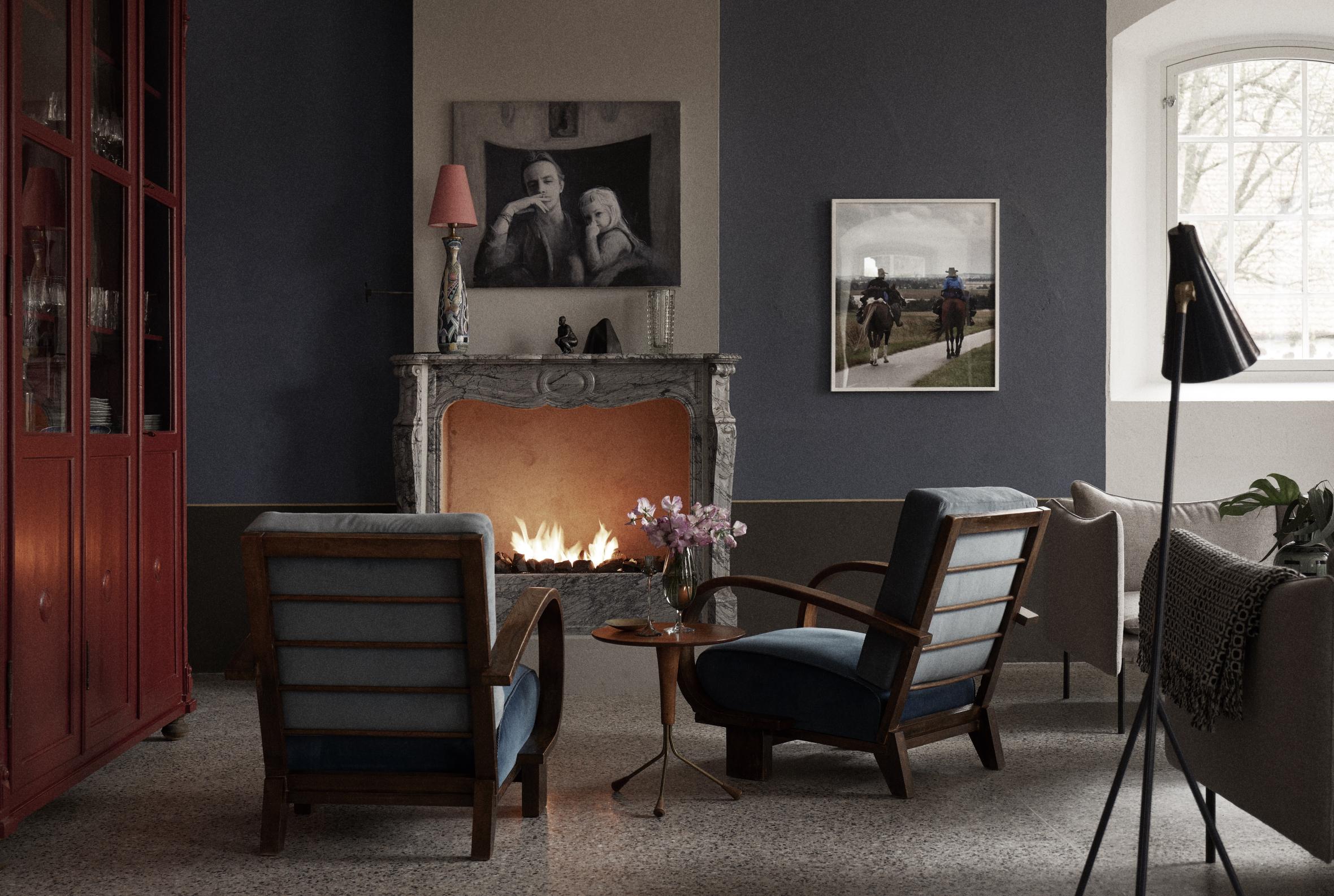 Elegant living room with lit marble fireplace, mid-century armchairs, framed photographs, display cabinet, and standing lamp creating warm, sophisticated atmosphere