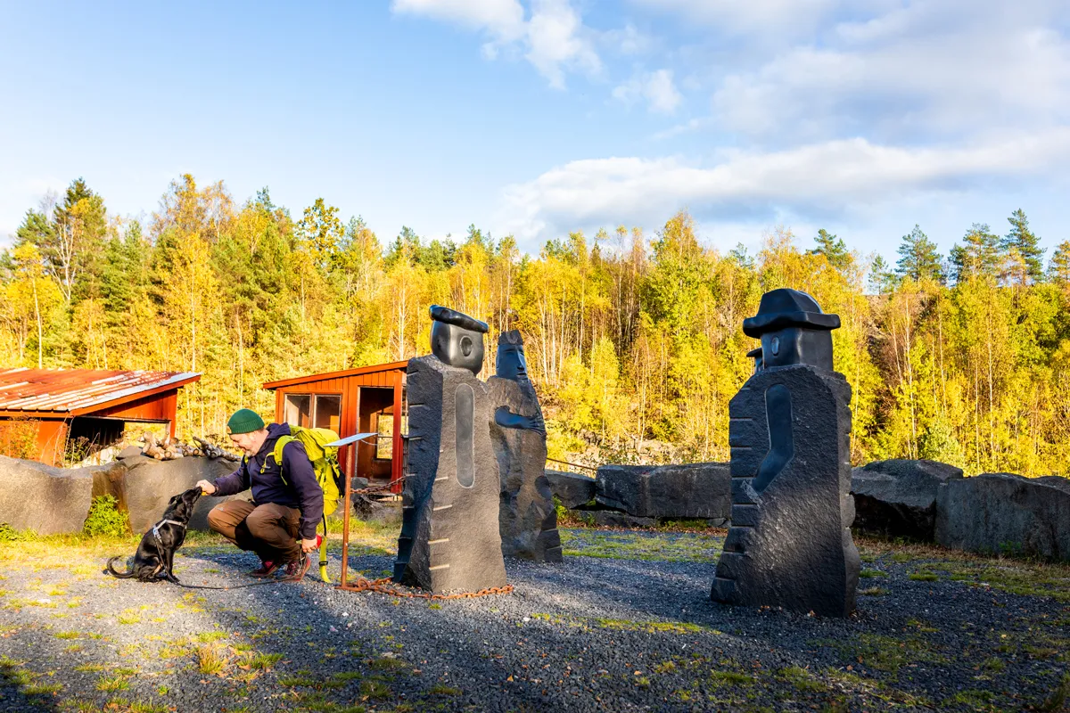 Man sitter på huk bredvid sin hund bredvid stora huggna stenskulpturer i stenbrottsmiljö, höstskogsbakgrund med gyllene björkar och klar himmel