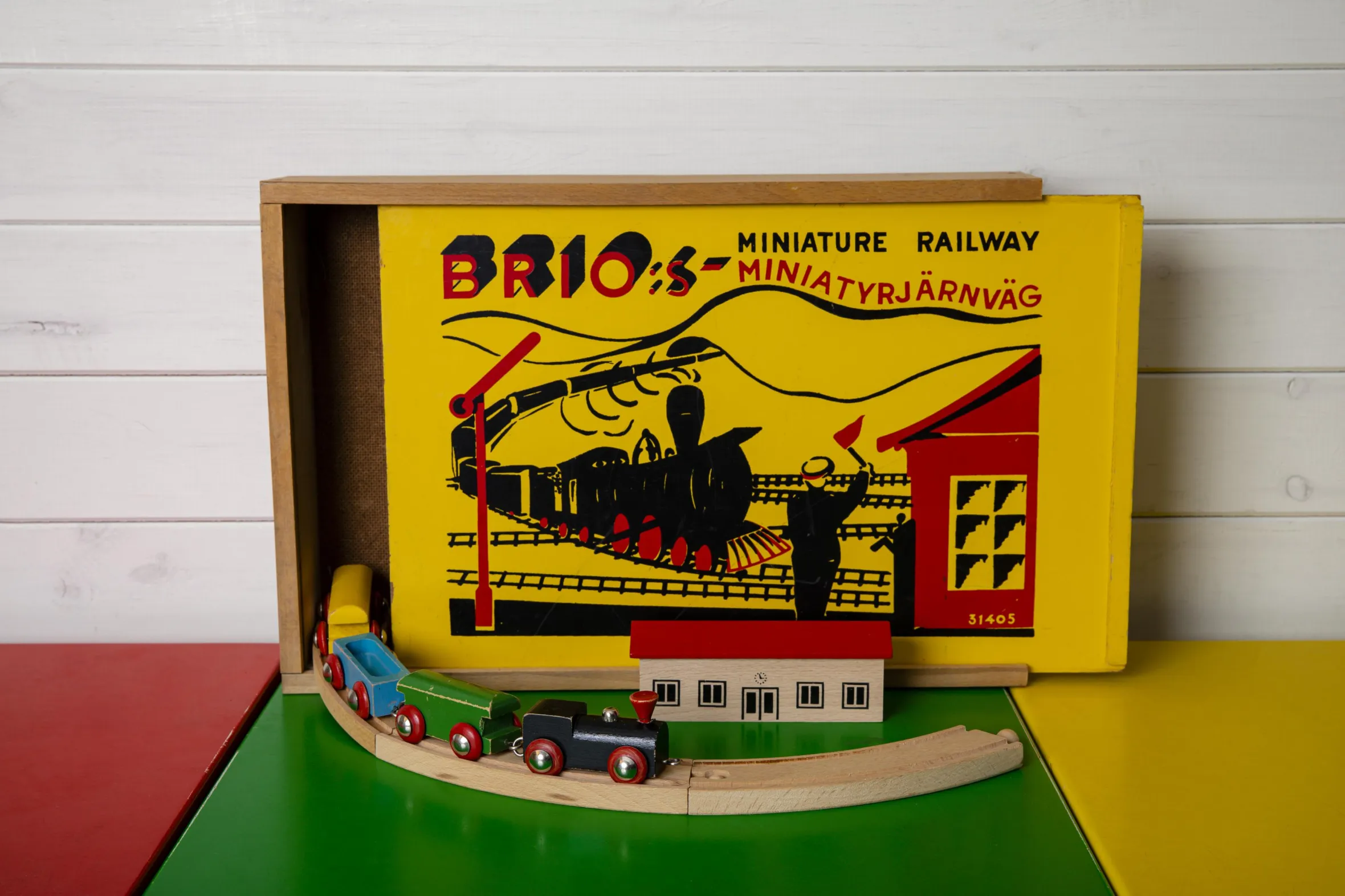 Vintage Brio Miniatureisenbahn-Spielset mit Originalverpackung, Holzzug und Schienen auf bunter Spielmatte vor weißer Holzwand ausgestellt