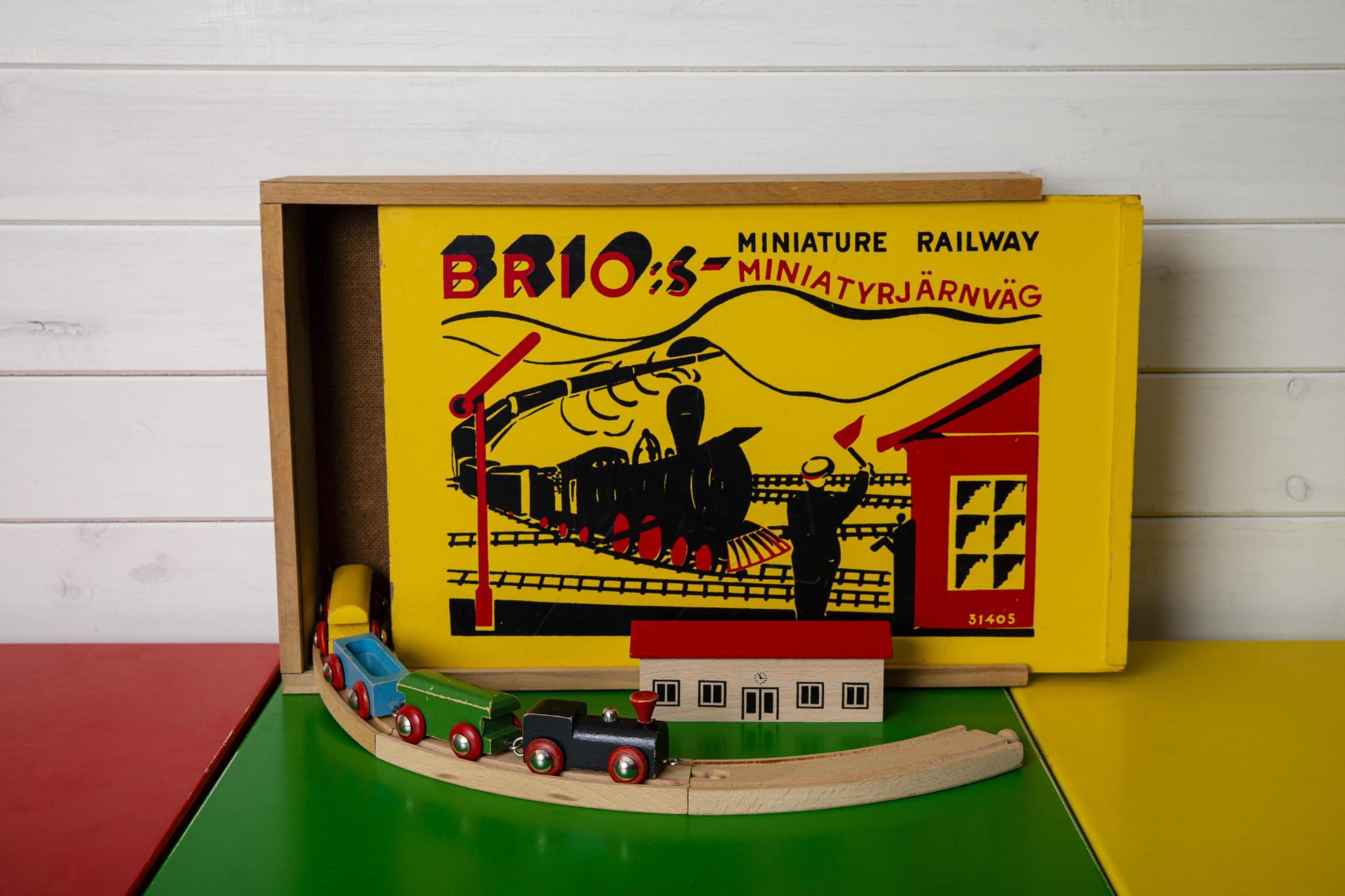 Vintage Brio Miniatureisenbahn-Spielset mit Originalverpackung, Holzzug und Schienen auf bunter Spielmatte vor weißer Holzwand ausgestellt
