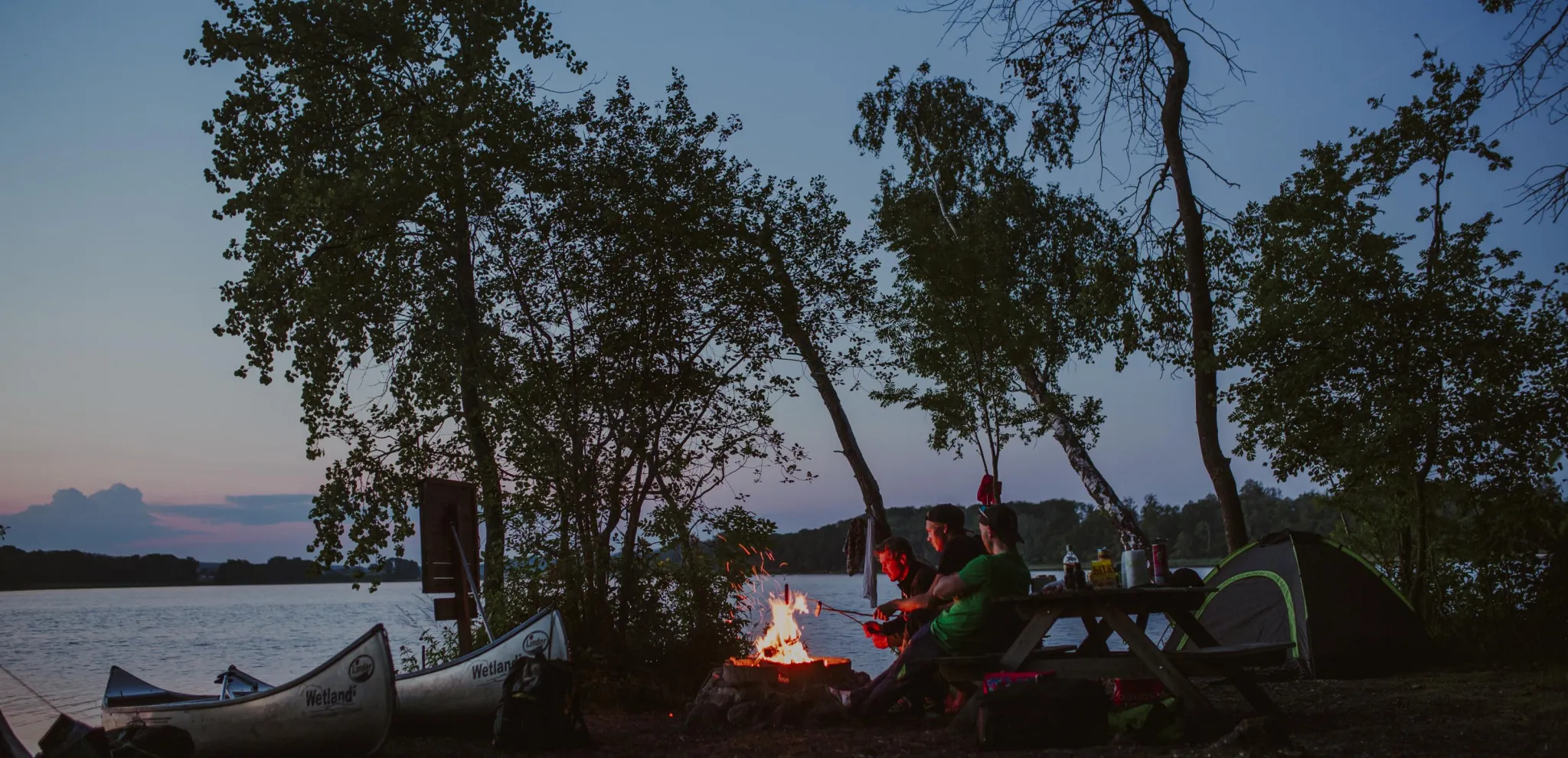 A group of friends grill hot dogs at dusk next to Lake Ivösjön