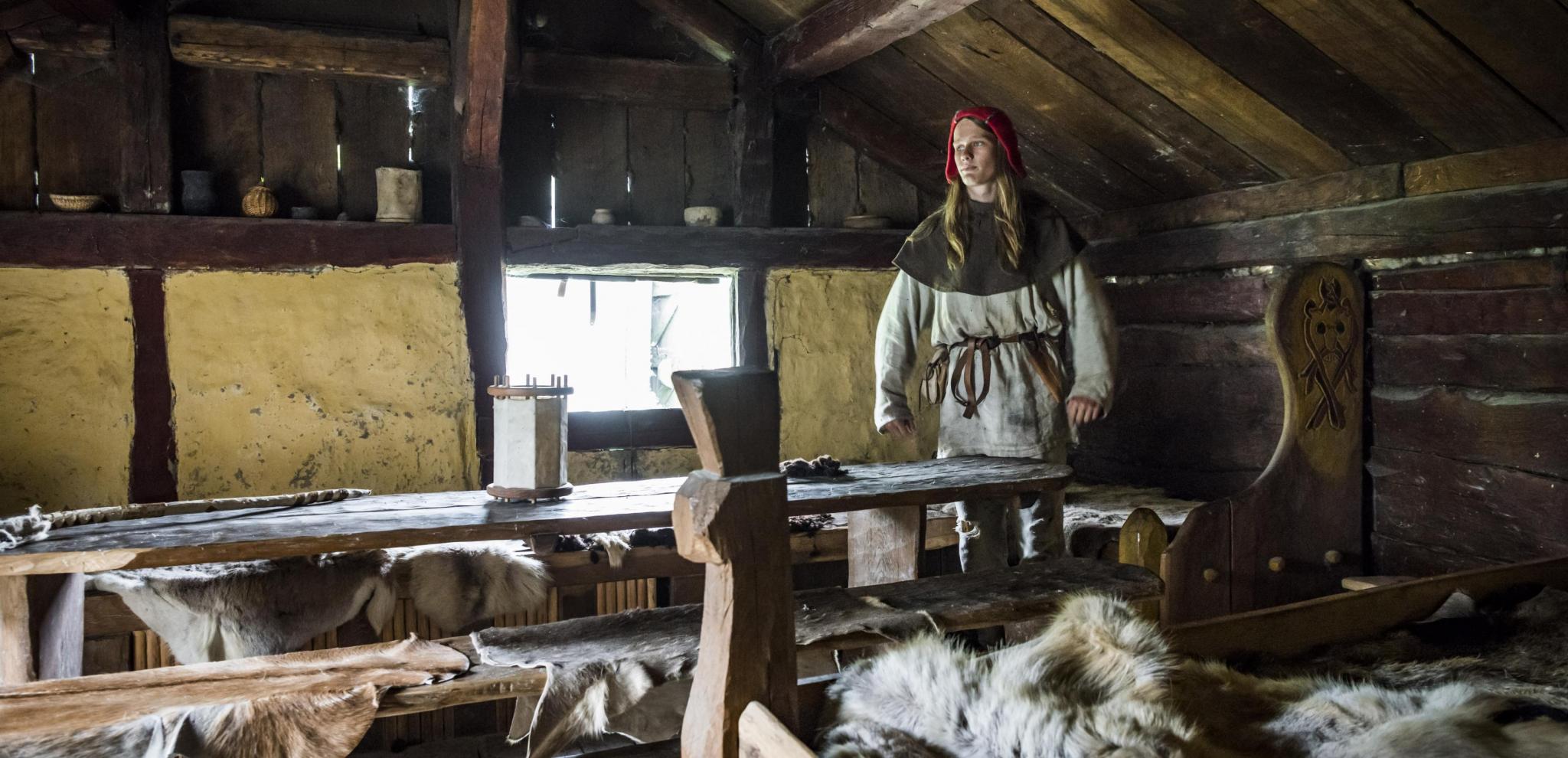 Ein junger, als Wikinger verkleideter Mann steht hinter Betten und einem langen Holztisch in einem nachgebauten Wohnhaus.