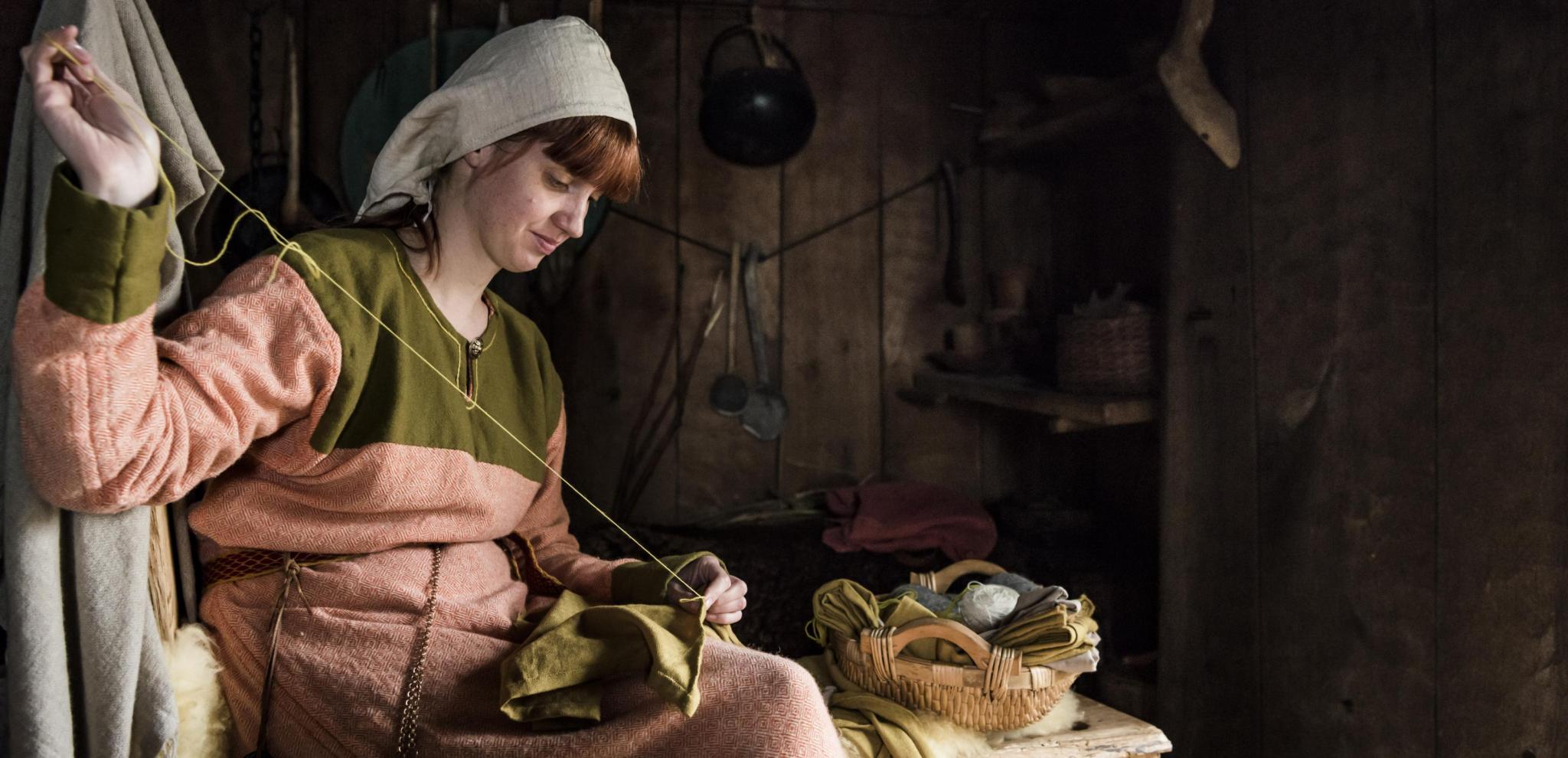 A viking woman sewing on a stool inside a woodhouse