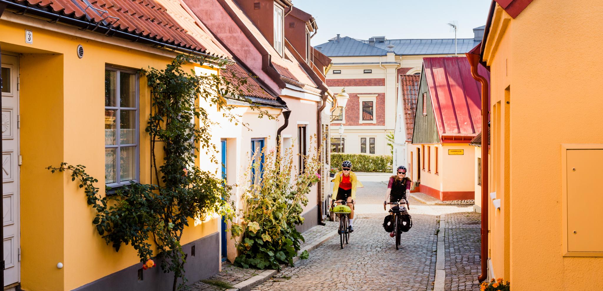 Two cyclists come cycling on a cobbled street among picturesque houses.