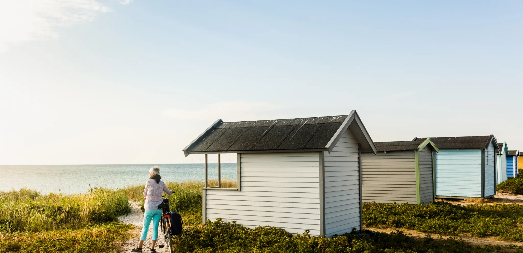 A woman with a bike standing next to a hut on the beach facing the sea 