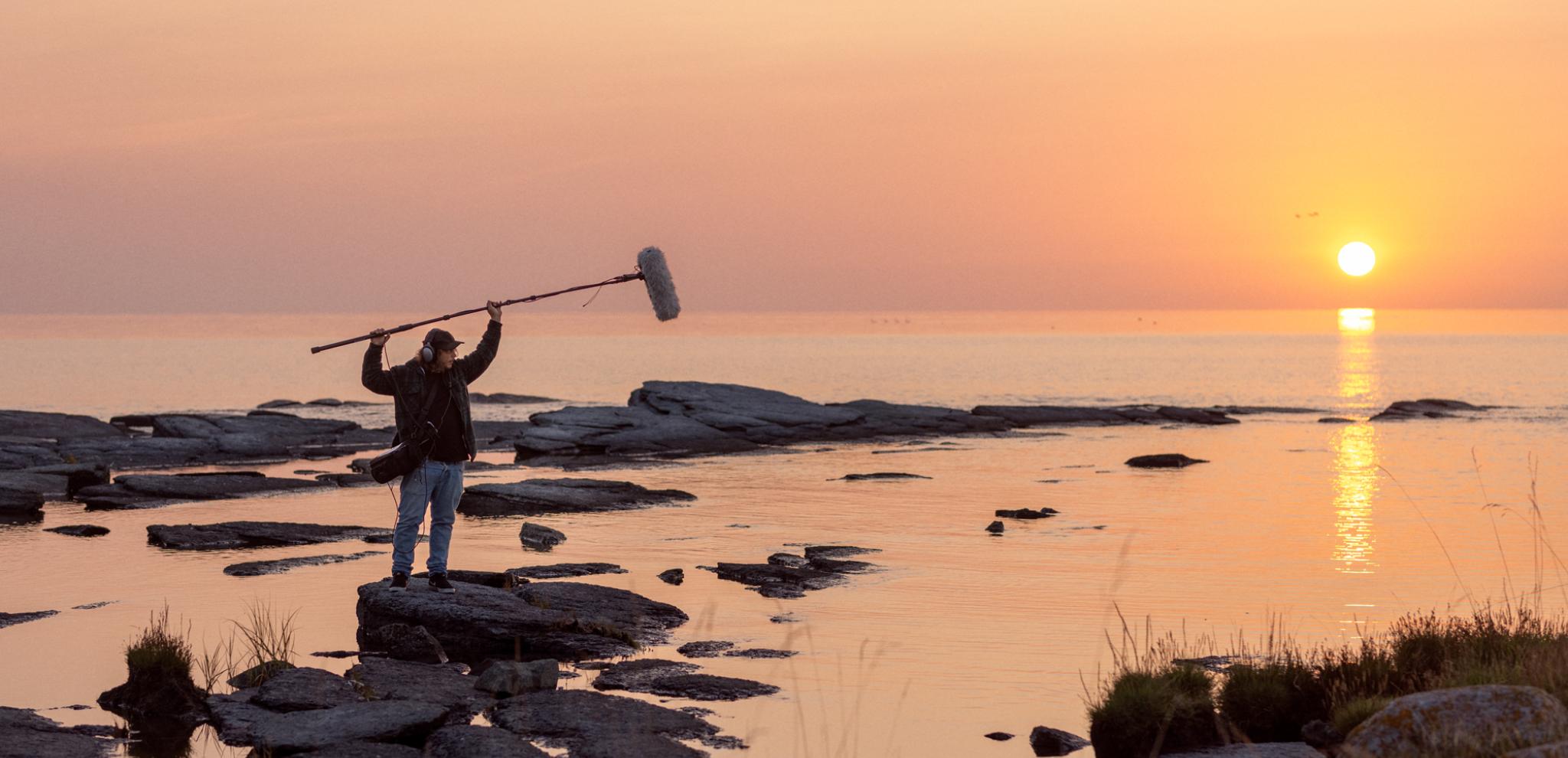 A person standing on rocks in sunrise holding a sound boom