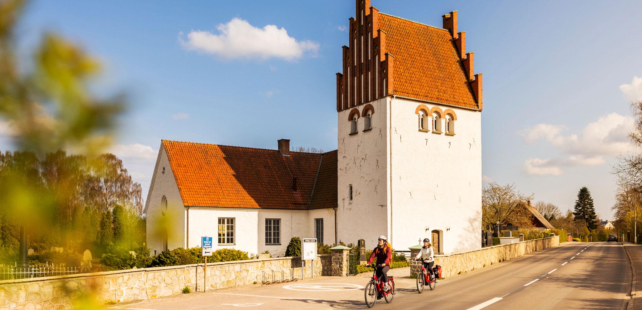 Medieval church with distinctive tower and stepped gable, two cyclists passing by on touring bikes along stone-walled road