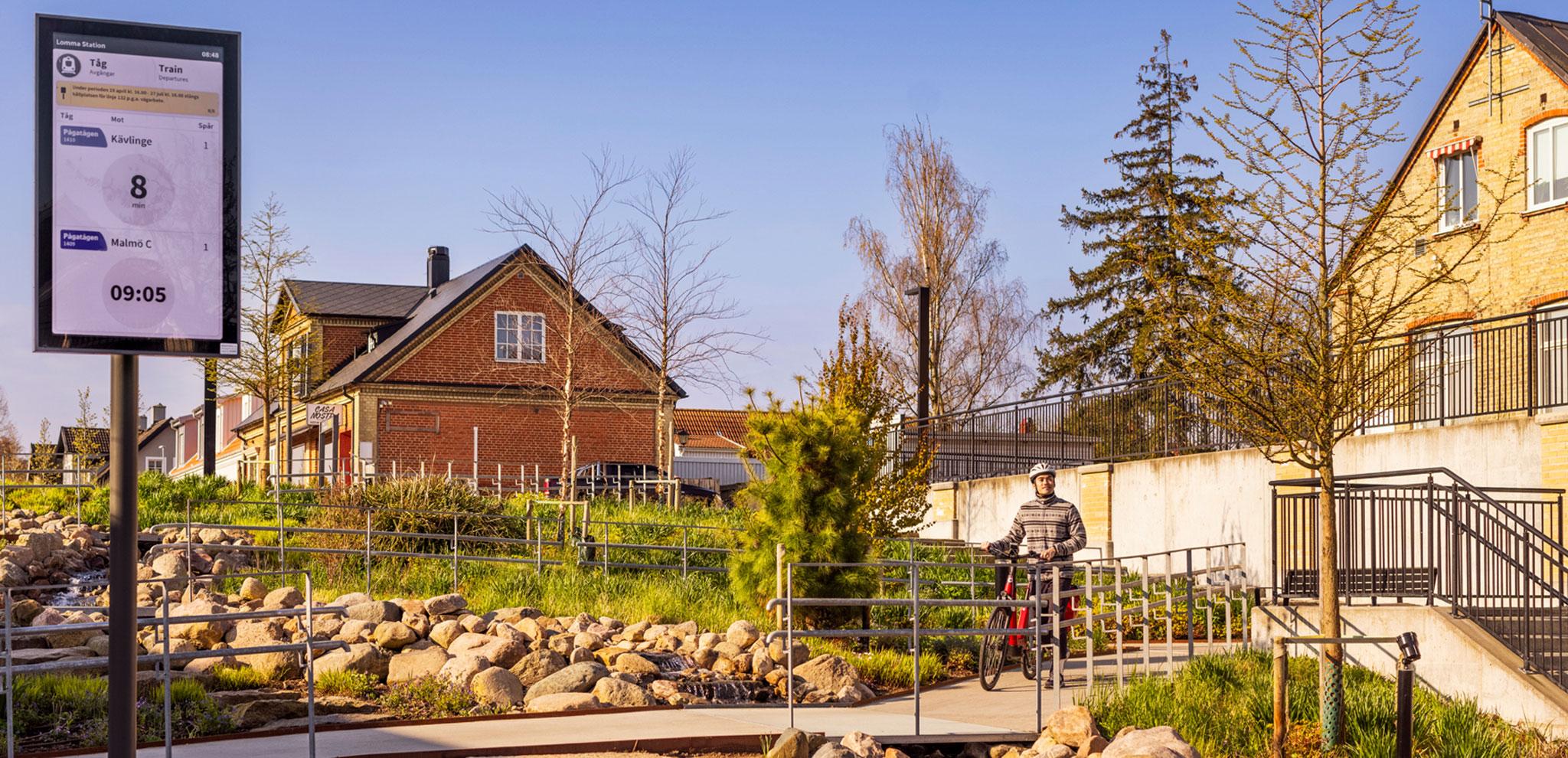 Train station with digital display showing 09:05 departure to Malmö C. Cyclist waiting by brick buildings and landscaped stone pathway