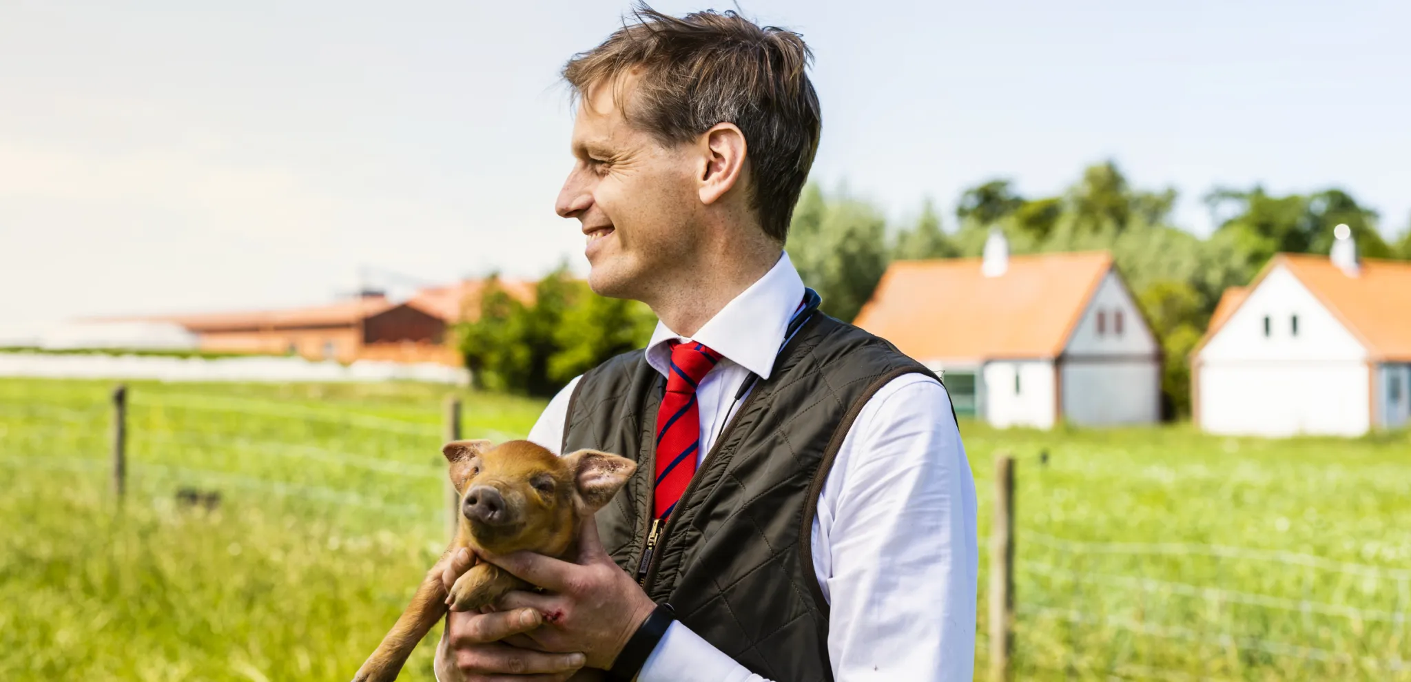 Smiling man in traditional vest and tie holding small pig outdoors with farm buildings and green fields in rural countryside background