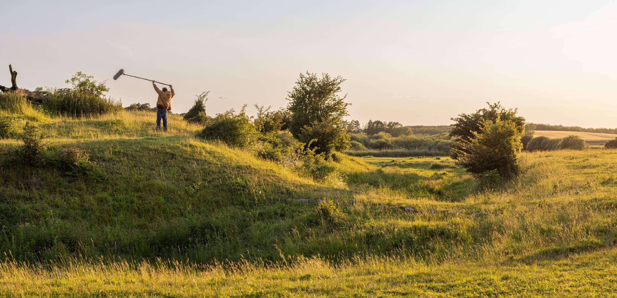 Sound guy standing on a grassy hill with  arms in the air with microphone.