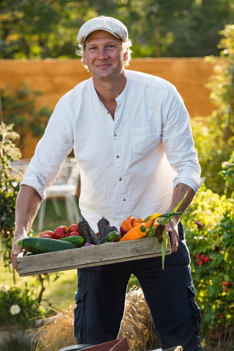 A man at Jord och bord standing in a green garden holding a tray of vegetables.