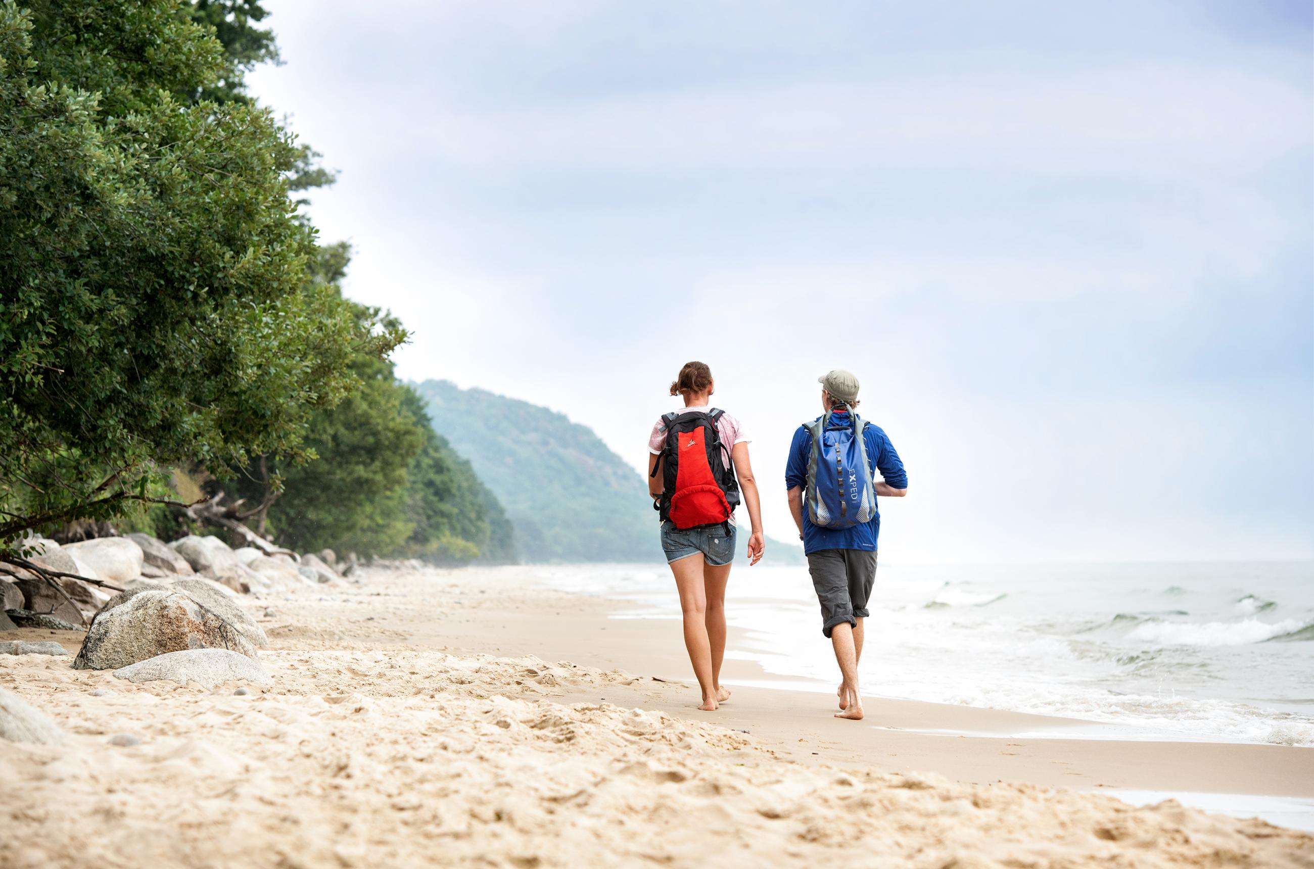 Ein Paar mit Wanderrucksäcken spaziert barfuß am Strand entlang, vorbei an einem Wald.
