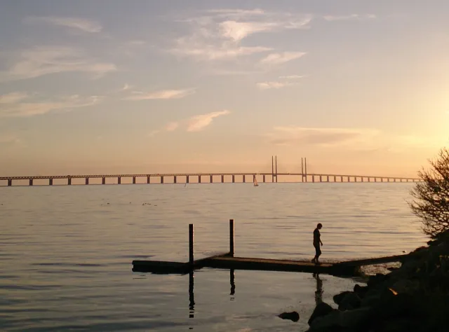 Eine Frau steht bei Sonnenuntergang auf einem Steg und hat die Öresundbrücke im Hintergrund.