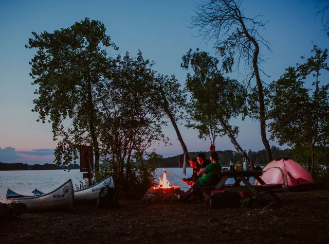 Drei junge Männer haben am See Ivösjön Zelte aufgebaut und grillen nach Sonnenuntergang Würstchen am Lagerfeuer.
