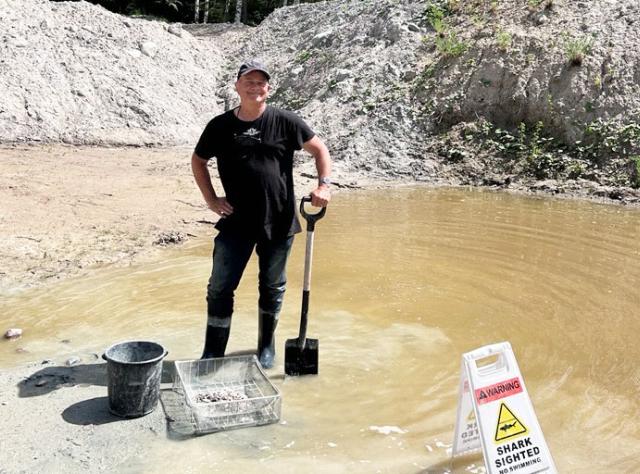 Person i grumligt vatten vid stenbrott med stövlar och siktutrustning intill humoristisk "SHARK SIGHTED NO SWIMMING"-skylt