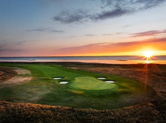 Golf green with sand bunkers overlooking water at sunset, sun creating dramatic rays across coastal links course with natural rough areas