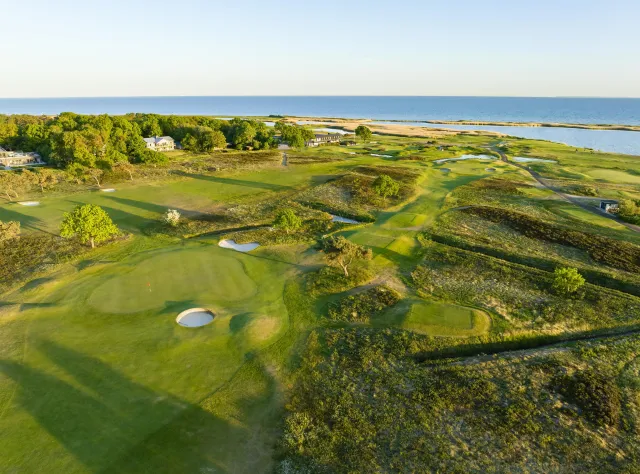 Flygvy över kustnära linksbana med böljande fairways, sandbunkrar, puttinggreener och hav i bakgrunden, hus inbäddade bland träden