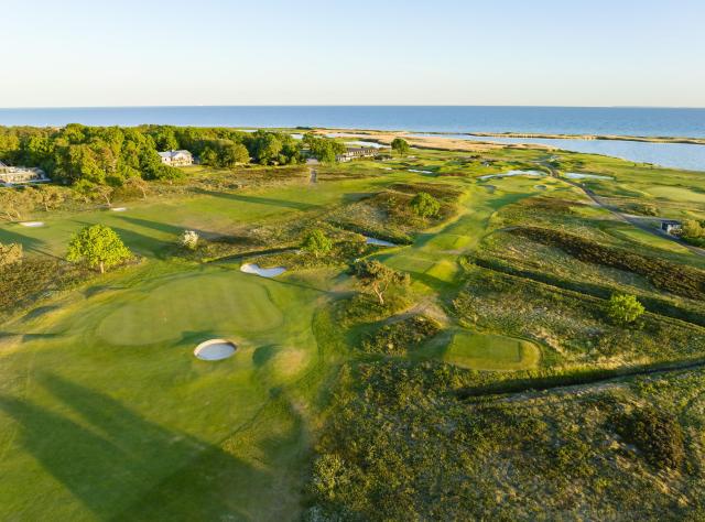 Flygvy över kustnära linksbana med böljande fairways, sandbunkrar, puttinggreener och hav i bakgrunden, hus inbäddade bland träden