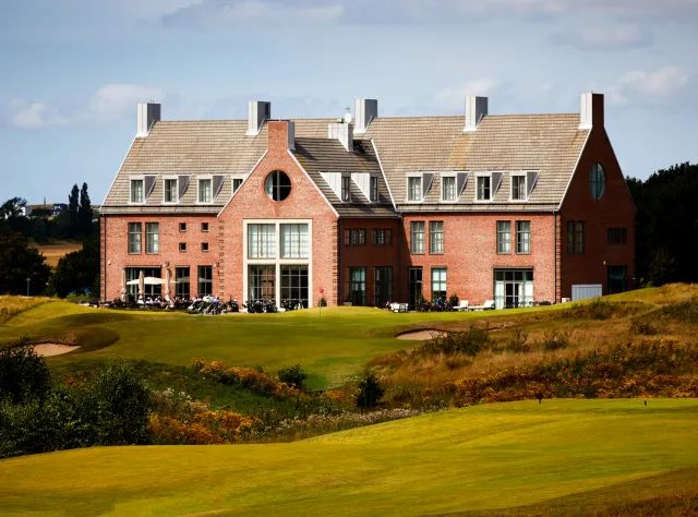 Stately brick clubhouse on manicured golf course with distinctive roof dormers, outdoor seating area, sand bunkers and rolling fairways in foreground