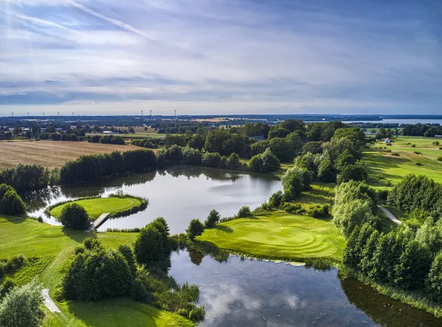 Aerial view of golf course with water hazards, tree-lined fairways and greens surrounded by lake, distant wind turbines and countryside landscape