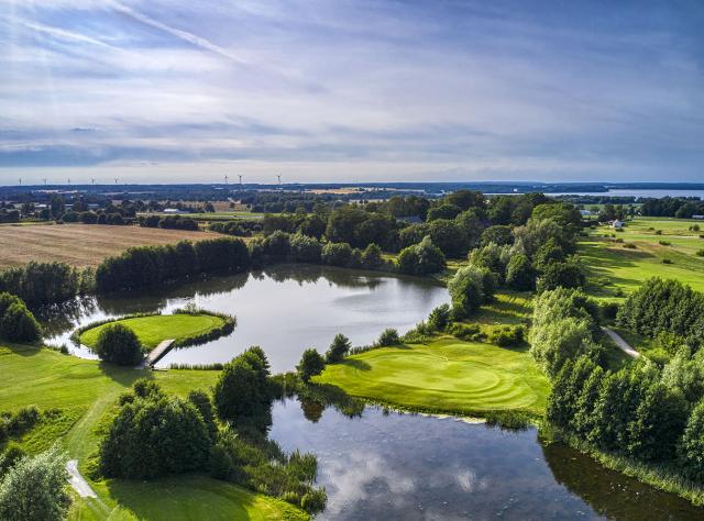 Flygvy över golfbana med vattenhinder, trädkantade fairways och greener omgivna av sjö, avlägsna vindkraftverk och landsbygdslandskap