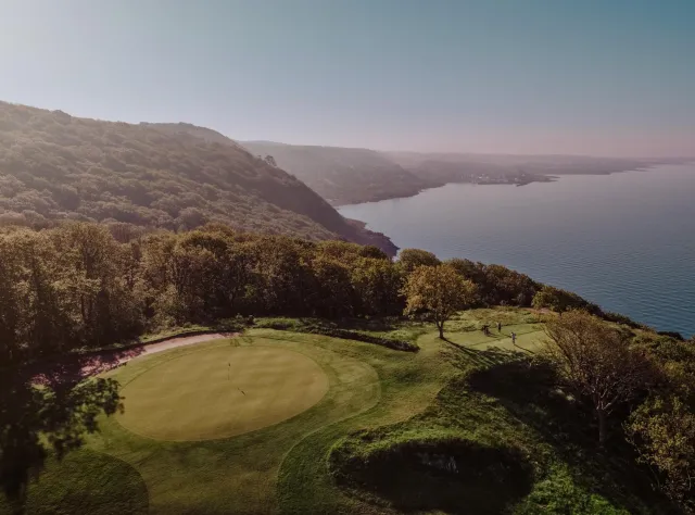 Elevated view of dramatic clifftop golf hole with putting green overlooking coastline, golfers playing amid wooded hillsides and expansive bay view