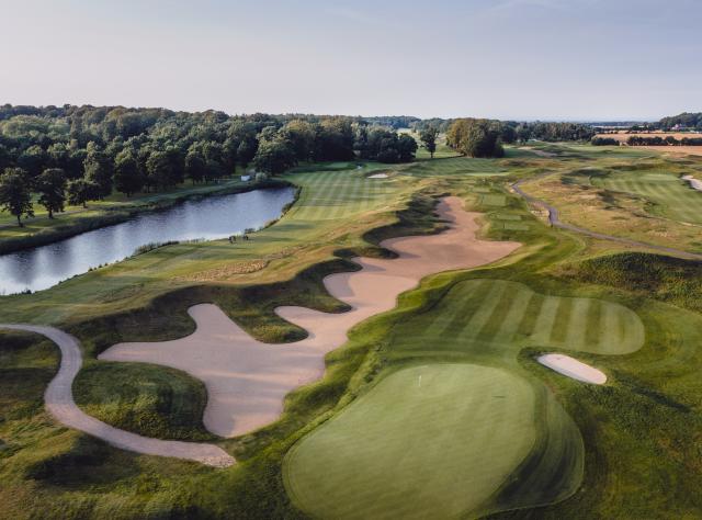 Aerial view of golf course featuring large sand bunkers around greens, water hazard lake, and dense forest backdrop