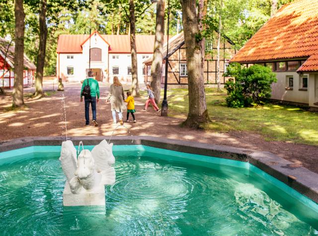 Dekorativ fontän med vattensprutande skulptur i förgrunden, familj som promenerar förbi historiska byggnader med traditionella tegeltak i museiby