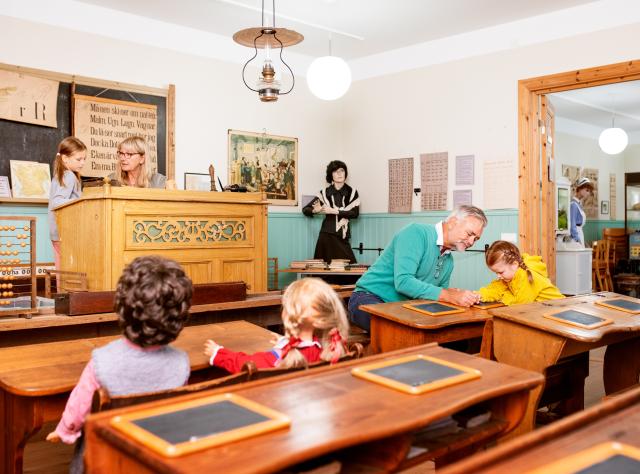 Historiskt klassrum med träbänkar och skifferplattor, en morfar hjälper barn att skriva, en mormor leker lärare vid utsmyckad träpulpet
