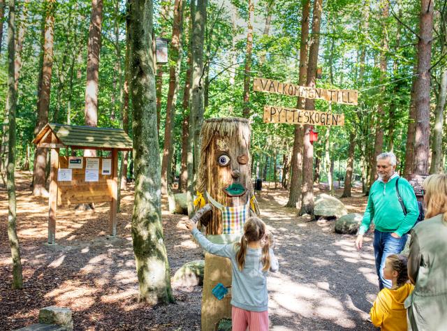 Familj som utforskar äventyrpark i skogen med träskulpturer, kojor och snidda trollfigurer