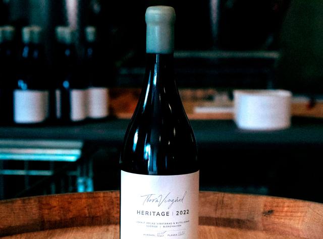 Wine bottle labeled "Thora Vingård Heritage 2022" on wooden surface in atmospheric winery cellar with blurred wine storage in background