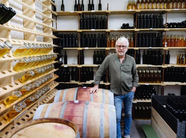 Winemaker with glasses standing beside wooden wine barrel in cellar, surrounded by shelves filled with wine bottles and wooden wine racks