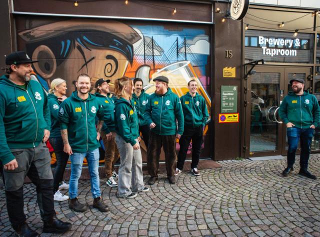 Grupp människor i matchande gröna huvtröjor som står utanför bryggeri med färgglad väggmålning, kullerstensgata och Brewery & Taproom-skylt synlig