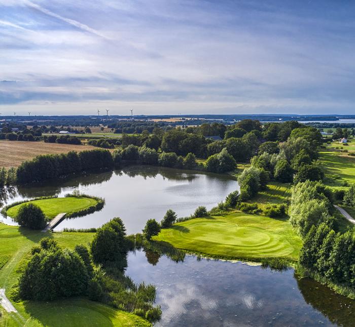 Aerial view of golf course with water hazards, tree-lined fairways and greens surrounded by lake, distant wind turbines and countryside landscape