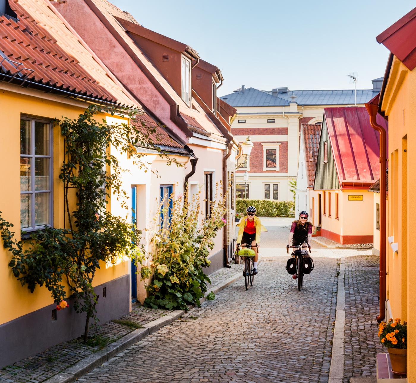 Two cyclists come cycling on a cobbled street among picturesque houses.