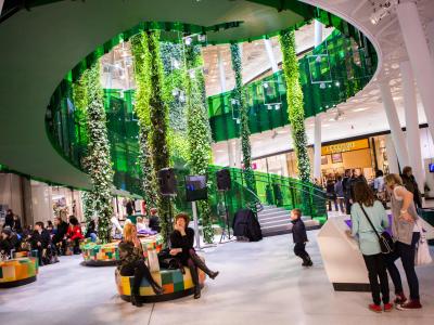 Eine auslandende Wendeltreppe im Einkaufszentrum Emporia Malmö, unter der sich Shopper auf Sitzgelegenheiten ausruhen.