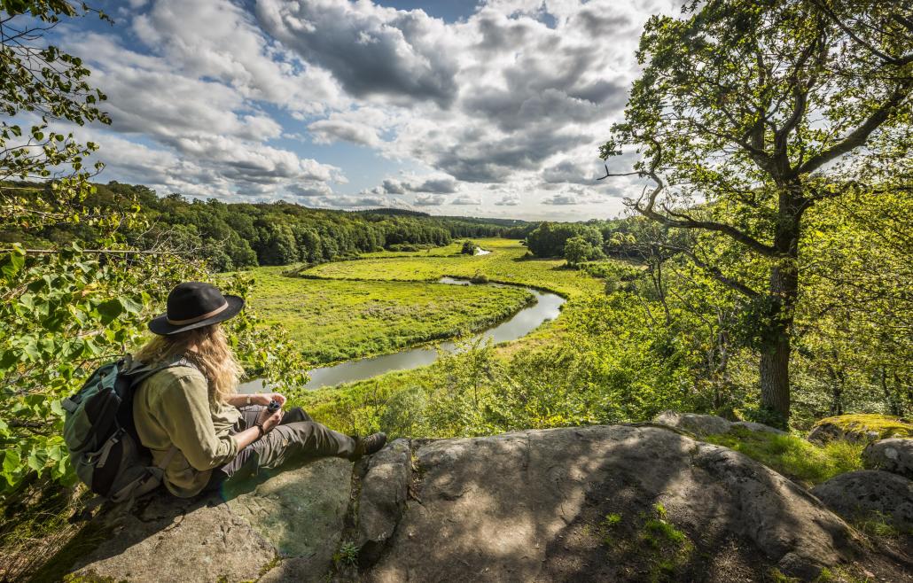 Eine Frau mit Hut sitzt auf einer Klippe und blickt auf ein grünes Flusstal.